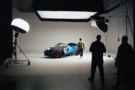 Im hell erleuchteten Studio umgibt ein Puma x BMW Rennwagen mit blauen und roten Grafiken drei Personen, die entspannt posieren. Große Scheinwerfer und Studioausrüstung setzen die Szene im dokumentarischen Stil in Szene. Fotografie von Alex Sobotta. Behind the scenes, race car, team, studio shoot, Alex Sobotta photo. Alex Sobotta Fotograf