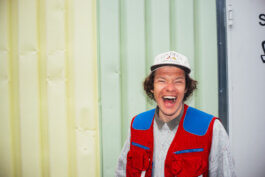 Dokumentarisches Porträt: Hagen Schäfer mit lockigem Haar, weißer Kappe, grauem Pullover und roter Weste mit blauen Details lacht gelöst vor einer hellgelb-grünen Wand. Fotografie von Alex Sobotta. The subject radiates joy in a colorful urban setting. Alex Sobotta Fotograf