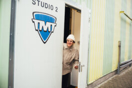 Fotografie von Alex Sobotta: Dex lächelt, schaut mit weißer Beanie und Brille filmreif durch die geöffnete Studio 2-Tür, markiert mit blauem TMT-Schild. Im Hintergrund lichtdurchflutete grün-gelbe Fassade. Photographer Alex Sobotta captured a joyful moment at the studio entrance. Alex Sobotta Fotograf