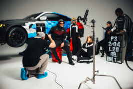 Fünf modisch gekleidete Menschen posieren vor einem BMW-Rennwagen im Studio, beleuchtet von weichem Licht, umgeben von Rennausrüstung und Schildern. Ausdrucksstarke Gesichter, authentische Posen. Fotografie von Alex Sobotta. Five people and a race car, motorsport-themed shoot. Alex Sobotta Fotograf