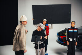 Im Studio neben einem schwarzen BMW stehen vier Personen. Ein großer schwarzer Reflektor wird gehalten, ein Monitor justiert. Eine Frau im Rennanzug und Marco Schöler beobachten konzentriert das Geschehen. Foto von Alex Sobotta. Puma x BMW photo shoot in progress, behind the scenes documentary style by Alex Sobotta. Alex Sobotta Fotograf