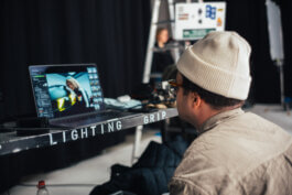 Eine Person mit weißer Mütze und Brille sitzt an einem Tisch mit „Lighting Grip“-Schild, vertieft in die Bildbearbeitung am Laptop. Im unscharfen Hintergrund eine Leiter und Silhouette. Foto von Alex Sobotta, Fotografie Alex Sobotta. Cinematic behind-the-scenes moment during Puma x BMW shoot. Photographer, lighting setup, and editing in focus. Alex Sobotta Fotograf