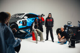 In einem Studio posiert eine Gruppe um einen blau-weißen Rennwagen, manche stehen, andere hocken; Werkzeug liegt griffbereit. Dramatisches Licht betont konzentrierte Gesichter—Fotografie von Alex Sobotta. Behind the scenes BMW x Puma. Team with race car and tools. Alex Sobotta Fotograf