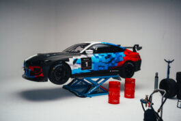 Im Studio leuchtet ein Rennauto mit pixelierten blauen, roten und schwarzen Grafiken erhöht auf einer Plattform. Rote Fässer, ein Reifen, Werkzeuge und Ausrüstung liegen rundherum. Fotografie von Alex Sobotta. Behind the scenes of a BMW race car shoot with Puma branding; workshop tools and props visible. Alex Sobotta Fotograf