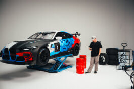 Szene im Studio: Ein Puma x BMW Rennwagen mit geometrischem Design schwebt auf einer Hebebühne. Ein Mann mit Basecap blickt aufs Handy, rote Fässer und Rennreifen im Hintergrund. Foto von Alex Sobotta Fotografie. Dynamic racing car in a studio setup. Alex Sobotta documentary style. Alex Sobotta Fotograf