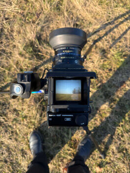 Cinematische Draufsicht auf eine Mamiya RZ67 Mittelformatkamera auf Stativ, durch den Sucher sieht man eine Landschaft. Umgeben von trockenen Gräsern und Schuhen einer Person. Fotograf: Alex Sobotta, Fotografie Stil. Photo by Alex Sobotta showing vintage gear and natural textures. Alex Sobotta Fotograf