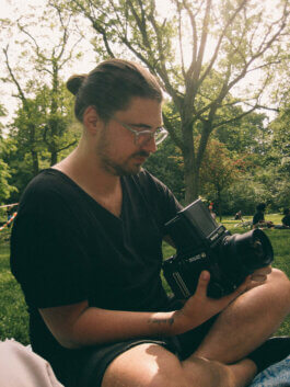 Nahaufnahme einer Person mit Brille und Dutt, die im Park im Schneidersitz auf dem Gras sitzt, konzentriert auf eine große Kamera in den Händen blickt. Im Hintergrund Bäume und entspannte Menschen. Fotografie von Alex Sobotta. A reflective photographer examines their camera outdoors. Photo by Alex Sobotta. Alex Sobotta Fotograf