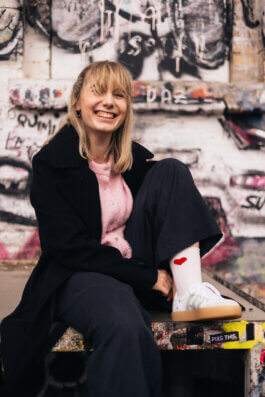 Lachende Elisabeth Becker sitzt vor einer mit Graffiti besprühten Wand, rosa Hemd unter schwarzem Mantel Alex Sobotta Fotograf