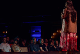 Jiaoying Summers steht auf der Bühne des Punchline Club Berlin im geblümten Kleid, im Vordergrund die Gesichter der ersten Publikumsreihe im blauen Bühnenlicht. Alex Sobotta Fotograf