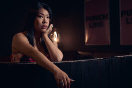Jiaoying Summers lehnt im Halbdunkel an der Bar des Punchline Club Berlin, den Kopf in die Hand gestützt, hinter ihr die beleuchteten Punchline-Schilder. Alex Sobotta Fotograf
