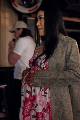 Jiaoying Summers im geblümten Kleid und olivfarbenem Mantel im Club, im Hintergrund ihr Mann im Gespräch mit einer weiteren Person. Alex Sobotta Fotograf