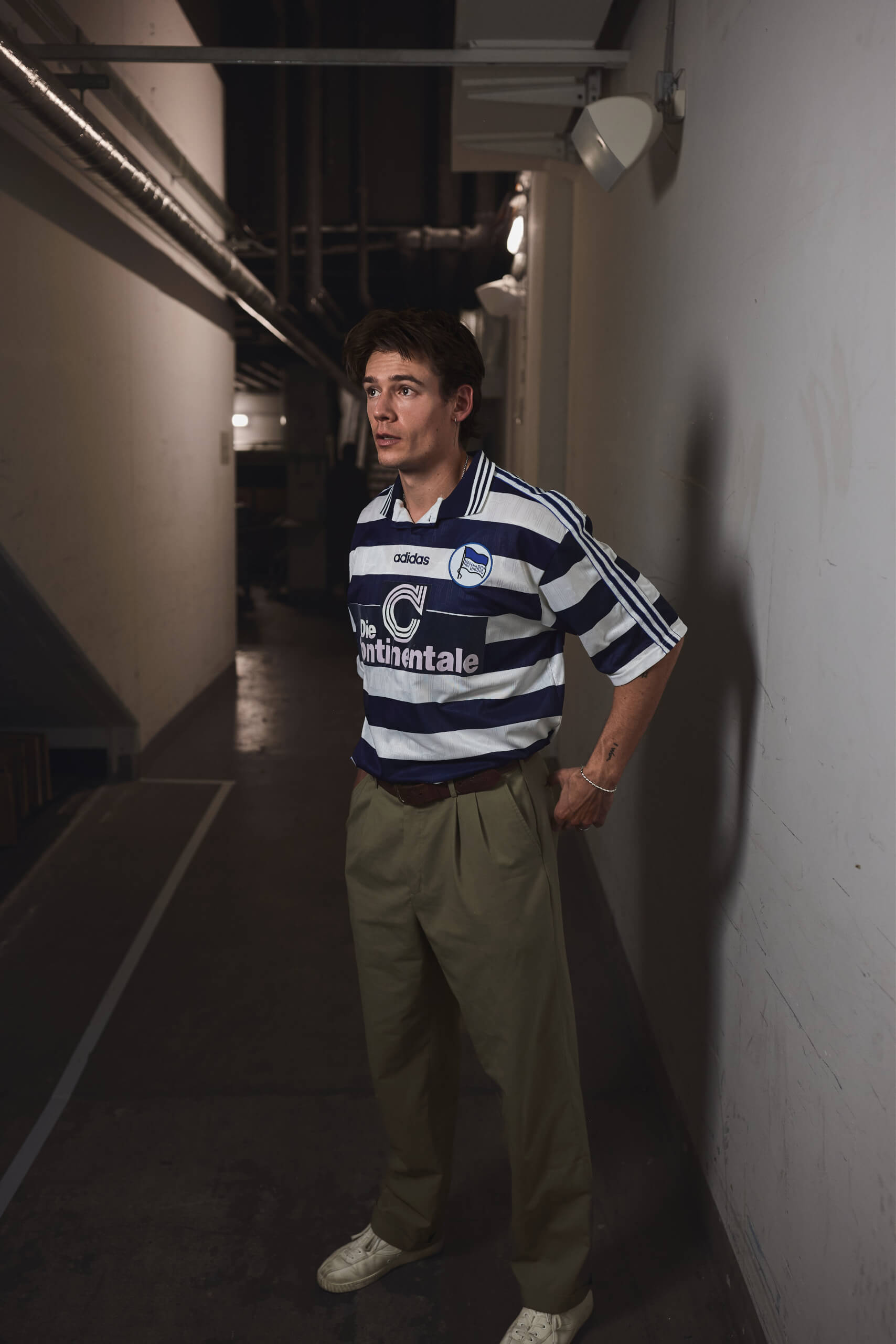 Mario Adrion in ganzer Figur in einem dunklen Gang, seitliches Licht auf dem Trikot, der Flur verliert sich hinter ihm im Schwarz. Alex Sobotta Fotograf