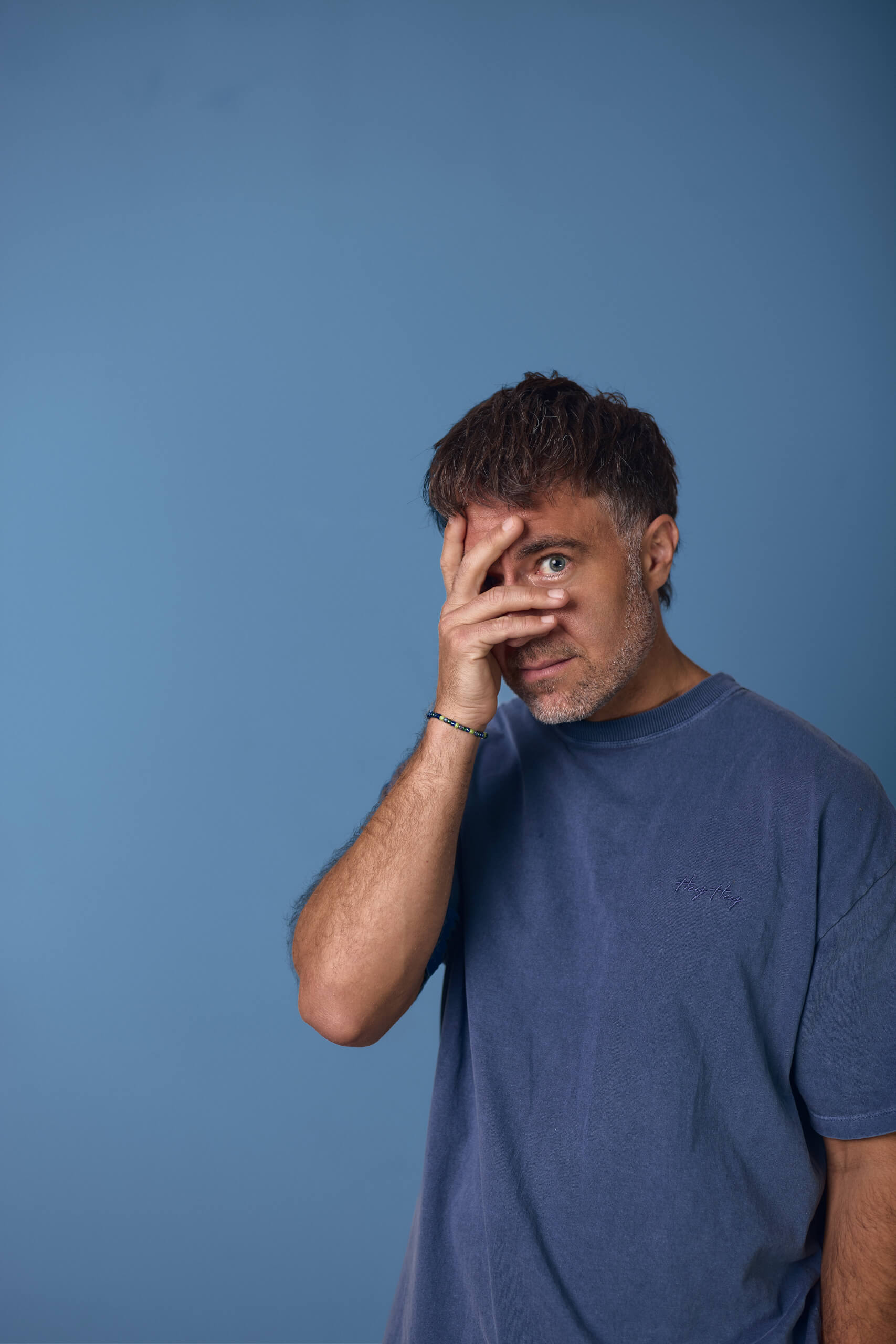 Moritz Hohl hält sich die Hand vors Gesicht und lugt mit einem Auge hervor, blaues T-Shirt vor blauem Hintergrund. Alex Sobotta Fotograf