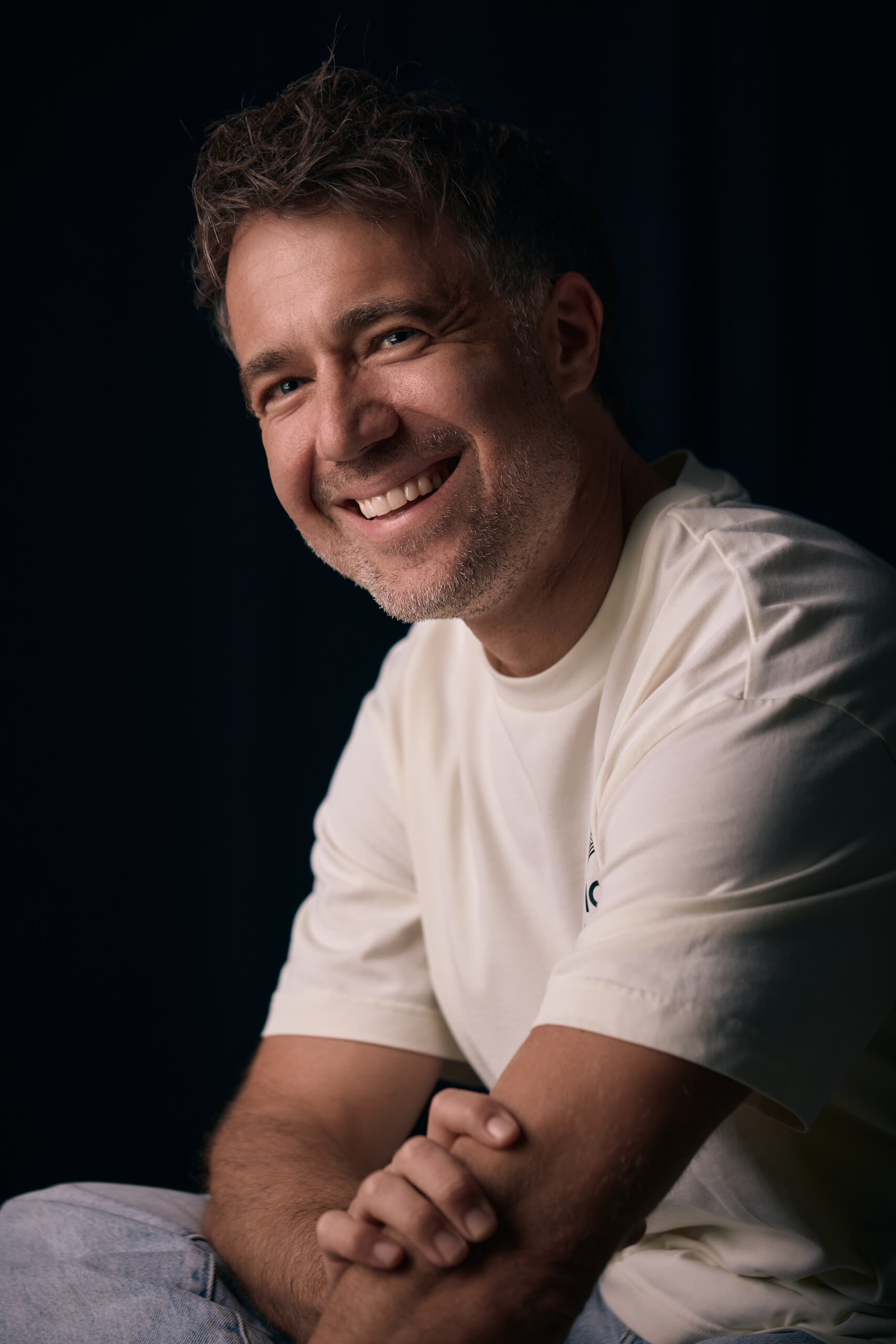 Moritz Hohl sitzt im weißen Shirt vor dunklem Hintergrund und lacht, die Hände ineinander gelegt. Alex Sobotta Fotograf