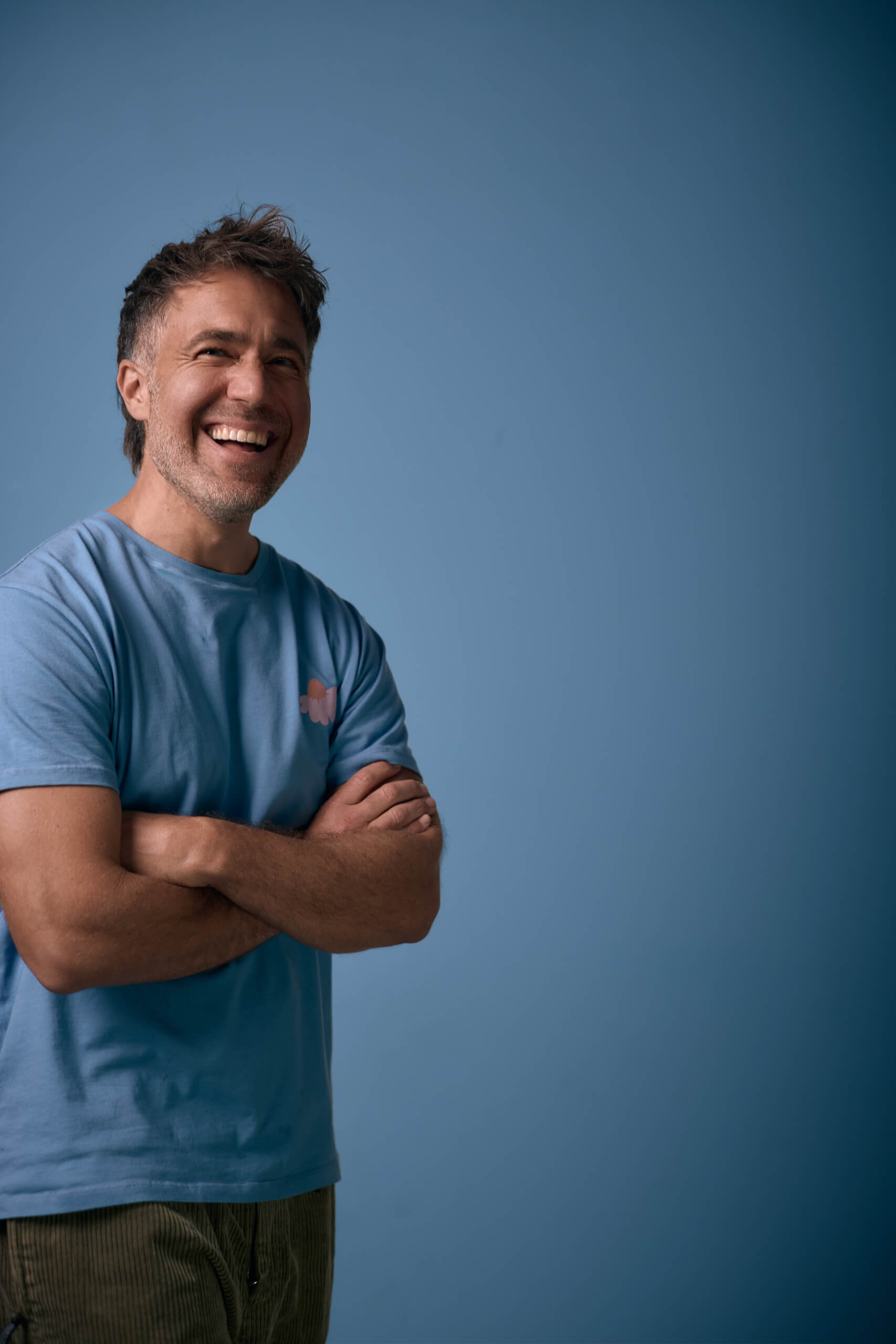 Moritz Hohl mit verschränkten Armen vor blauem Hintergrund, warmes Lachen, blaues T-Shirt. Alex Sobotta Fotograf
